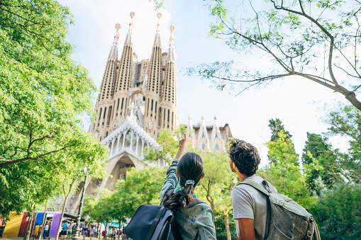 Rear view of male and female vacationers with backpacks looking up and pointing at distinctive towers of Sagrada Familia in Barcelona.