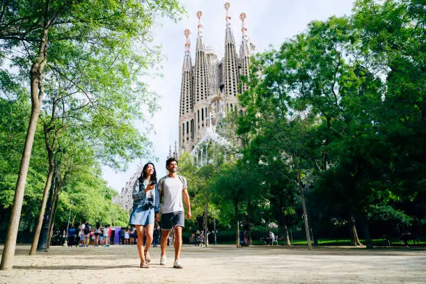 Mid distance low angle view of male and female tourists in 20s and 30s holding hands and walking through Barcelona park near Sagrada Familia.