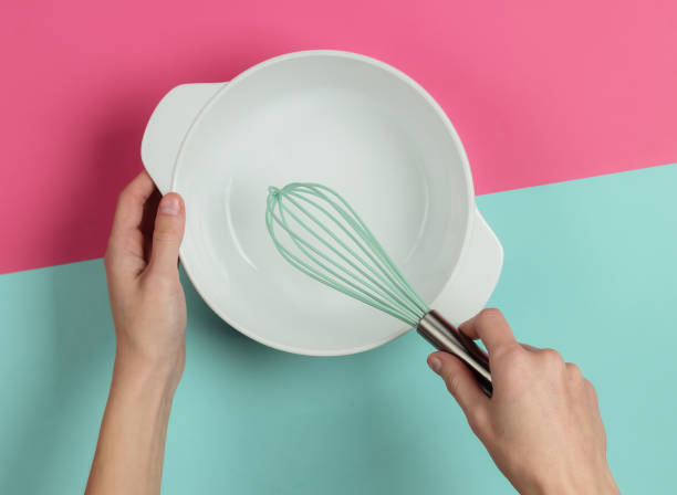 istock Female hands holding empty ceramic bowl and whisk on pink blue background. Top view 1194271998