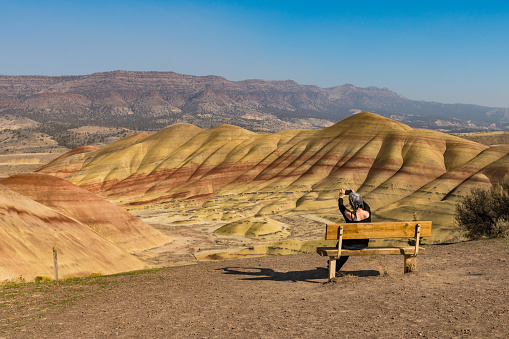 A young brunette with gray strands takes photographs with her smartphone sitting on a bench in Painted Hills Overlook, Oregon, USA.
