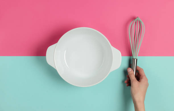 istock Female hands holding whisk on pink blue background with empty ceramic bowl. Top view 1194273612