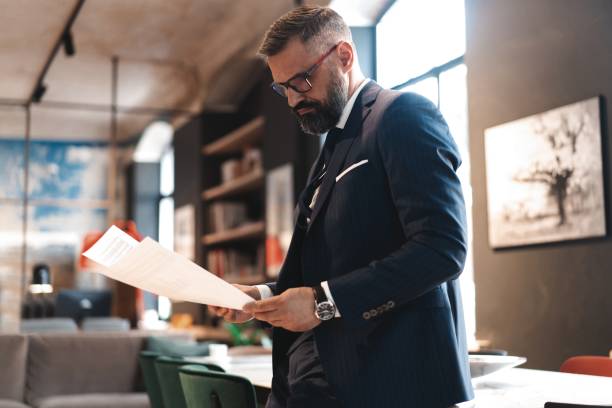 elegante hombre de negocios de pie cerca de la mesa en la oficina y la lectura de documentos - manager marketing recruitment small business fotografías e imágenes de stock