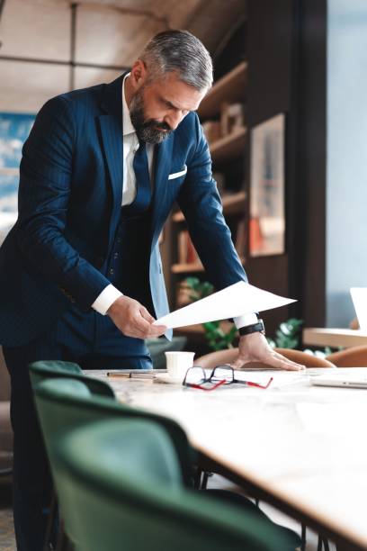 elegante hombre de negocios de pie cerca de la mesa en la oficina y la lectura de documentos - manager marketing recruitment small business fotografías e imágenes de stock