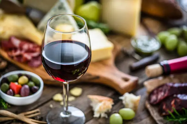 High angle view of delicious appetizer shot on rustic wooden table. The composition includes a selection of cheeses, Iberico ham, Spanish chorizo, red wine, grapes, olives and bread. A red wineglass is on foreground and the cheese board is out of focus at background. Predominant colors are red, yellow and brown. XXXL 42Mp studio photo taken with Sony A7rii and Sony FE 90mm f2.8 macro G OSS lens