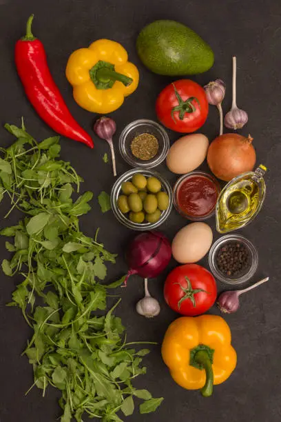 Photo of Rocket and spinach, eggs and tomatoes, yellow and red peppers, garlic, olives on a black background. Top view