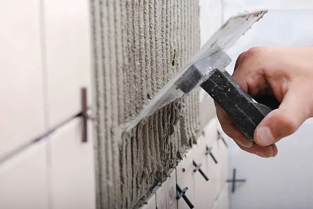 Closeup of man applying ceramic tile to a wall, working with trowel, putty kniife and mortar. Selective focus