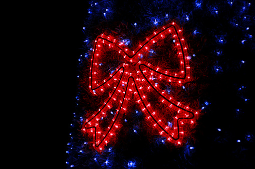 Part of Christmas tree decorations blue flashing lights. Detail of New Year string rice lights bulbs simulating shape of red decorative sparkle bows on street tree. Ornaments to christmas celebration.