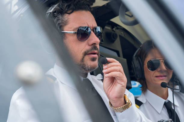 istock Black-haired man with a beard of approximately 40 years dressed in his pilot uniform accompanied by a blue helicopter cockpit, his co-pilot Latin woman with short blond hair and pilot uniform is soon to take off 1194308080