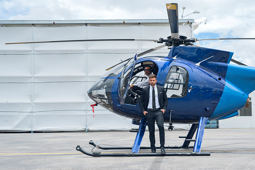 The Latin man of the executive co-pilot in a beautiful blue helicopter opens one of its doors to get rid and stands outside the helicopter and smiles dressed in a formal suit and next to the official pilot of the helicopter.