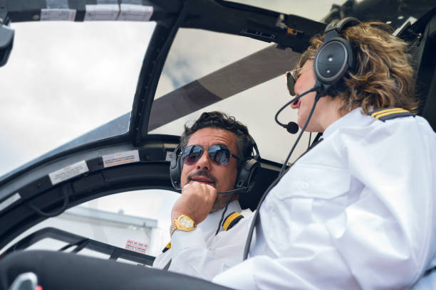 istock Black-haired man with a beard of approximately 40 years dressed in his pilot uniform accompanied by a blue helicopter cockpit, his co-pilot Latin woman with short blond hair and pilot uniform is soon to take off 1194308322