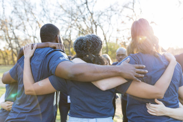 Rear view of united group of volunteers Group of diverse neighbors are unified while participating in a community cleanup event. They are standing with their arms around one another. charitable foundation photos stock pictures, royalty-free photos & images
