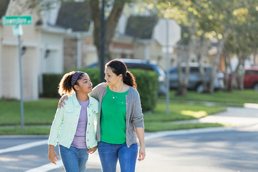 A mature Hispanic woman, in her 40s, walking with her mixed race Hispanic and African-American 11 year old daughter outdoors in their residential neighborhood. Her arm is around her the girl's shoulders as they cross a street. They are looking at each other with serious expressions. Perhaps mom is giving her daughter a pep talk or offering some motherly advice.