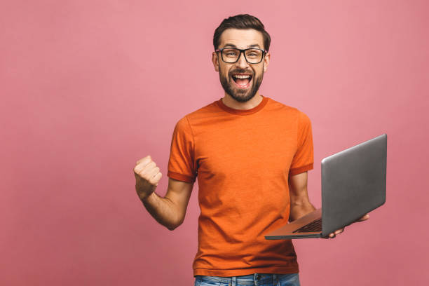 노트북 컴퓨터를 사용하여 분홍색 배경 벽에 고립 된 포즈를 취하는 행복한 흥분 한 젊은이의 이미지가 승자 제스처를 만듭니다. - businessman isolated laptop men 뉴스 사진 이미지
