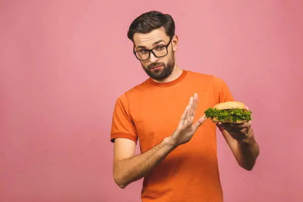 Photo of Handsome young man refusing unhealthy burger isolated over pink background. Diet concept. With copy space for text.