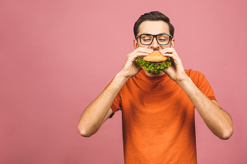 Young man holding a piece of hamburger. Student eats fast food. Burger is not helpful food. Very hungry guy. Diet concept. Isolated over pink background.
