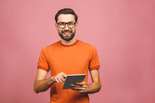 istock Happy young man in casual standing and using tablet isolated over pink background. 1194324095