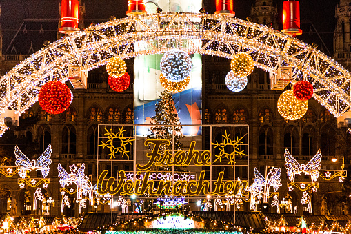 Color image depicting crowds of people on the street at one of the many Christmas markets in Vienna, Austria. The illuminated lights of the Christmas market can be seen beyond.