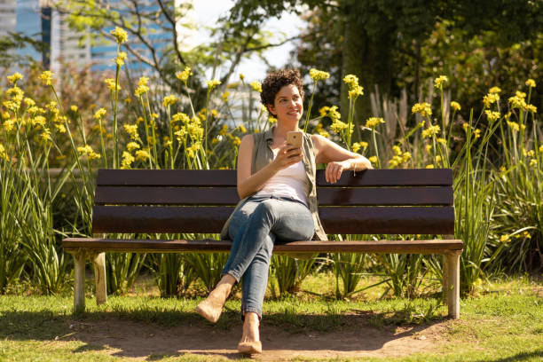 belle femme s'asseyant sur le banc de place publique parlant sur le téléphone portable - park bench photos et images de collection