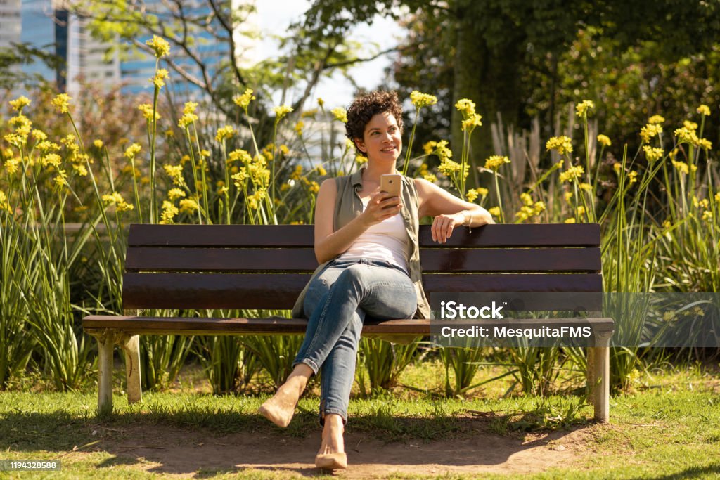 Beautiful woman sitting on public square bench talking on cellphone Public Park, Enjoyment, Nature, Wireless Technology, Using Phone Park Bench Stock Photo