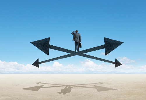 A businessman stands at a crossroad and the intersection of two arrows pointing in opposite directions as he considers what direction to go in.