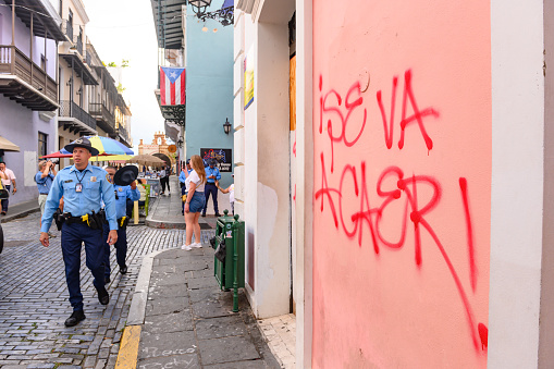 In Old San Juan, Puerto Rico uniformed officers patrol the area as tourists take photos. The wall is spray painted with an expression of protest.