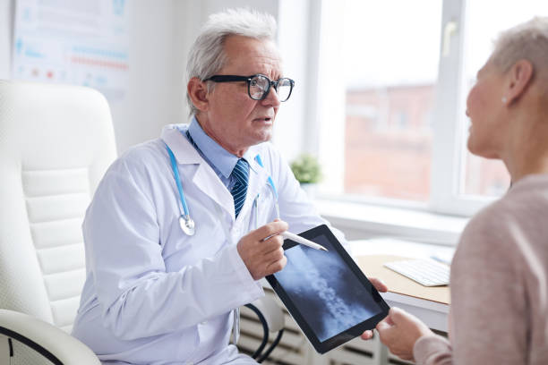 istock Confident senior male doctor in lab coat using digital tablet while telling patient about spinal cord disorder 1194338428