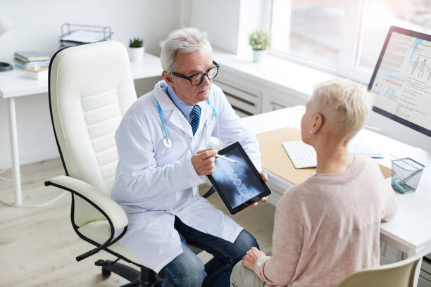 istock Serious aged doctor in white coat sitting at table and using tablet while showing online x-ray image of spine to patient 1194338442