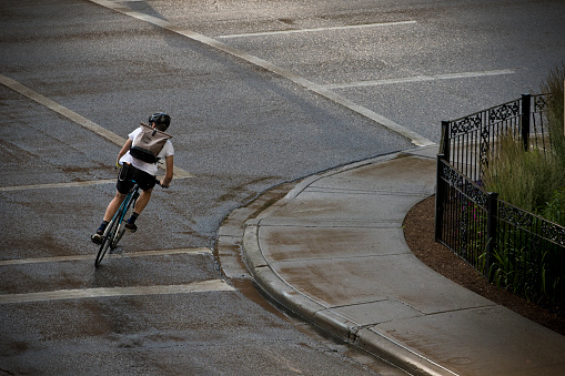 A male bicycle messenger rides a city street after a rainstorm. He is riding a fixed-gear style bicycle with a front brake and is wearing a bike courier backpack. He carries his bicycle lock in his belt and the key on his wrist.