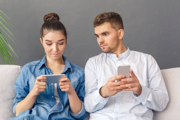 istock Relationnship Concept. Young couple sitting on sofa studio isolated on grey man with smartphone looking at woman watching video concentrated 1194372449