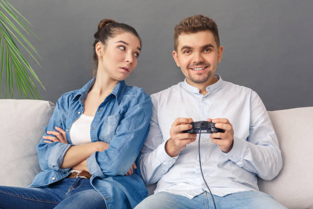 Relationnship Concept. Young couple man smiling excited playing video game sitting on sofa studio isolated on grey woman looking at him irritated
