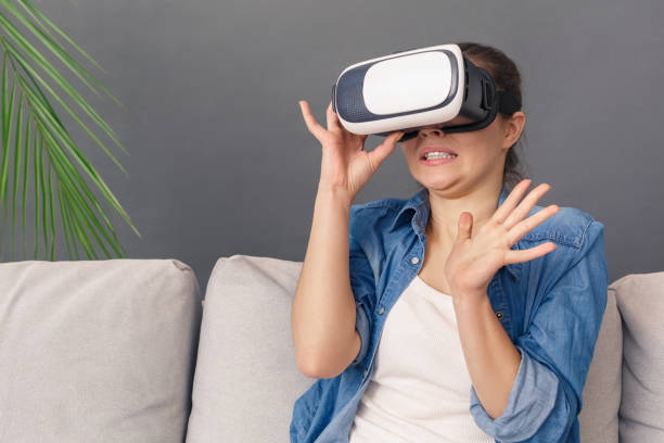 istock Freestyle. Young woman in virtual reality headset sitting on sofa studio isolated on grey watching video gesturing scared 1194372460