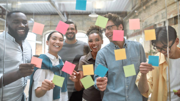 Working on business project together. Group of young and positive coworkers putting colorful sticky notes on a glass window in the creative office stock photo