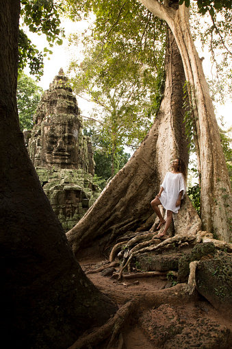 Young solo traveller, with brown long hair, wearing a white dress, walking between the root of giant tropical trees in the rainforest of Angkor Wat. In the background, a human face carved in stone.