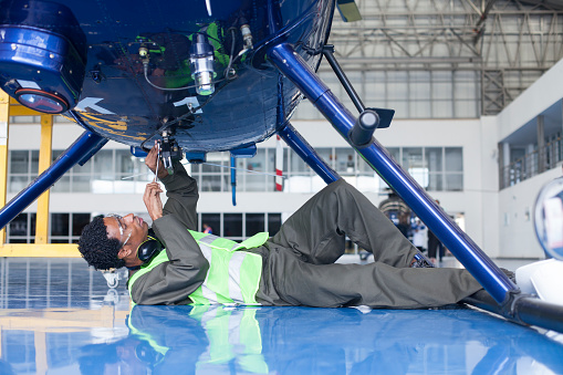 Latin man with curly hair and brown skin dressed in green work overalls reflective vest, and safety headphones resides very well from the bottom one of the helicopters stationed in the hangar where he works as a technician