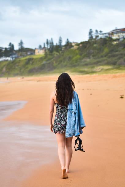Woman Walks Home Along Sandy Beach with Shoes in Hand A young woman walking the beach with no shoes after rainfall. walking beach women sadness stock pictures, royalty-free photos & images
