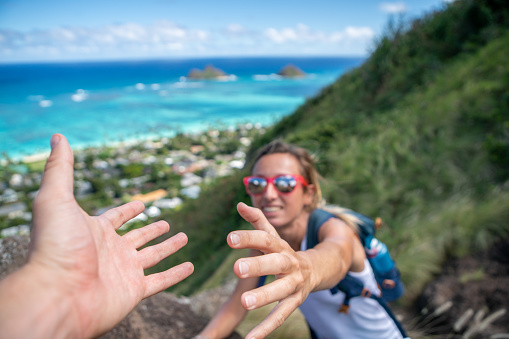 Hiker assisting teammate at mountain top giving a helping hand to reach summit; man helps woman moving up on trail above sea in Hawaii, USA