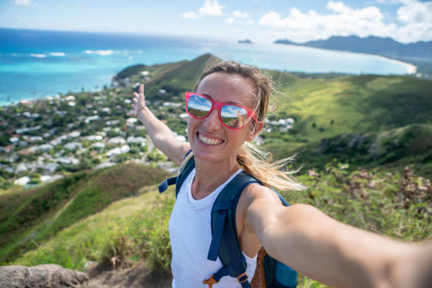 Young woman hiking in Hawaii takes selfie Young woman on mountain top overlooking the ocean taking selfie portrait using mobile phone, Oahu, Hawaii, USA reach destination stock pictures, royalty-free photos & images