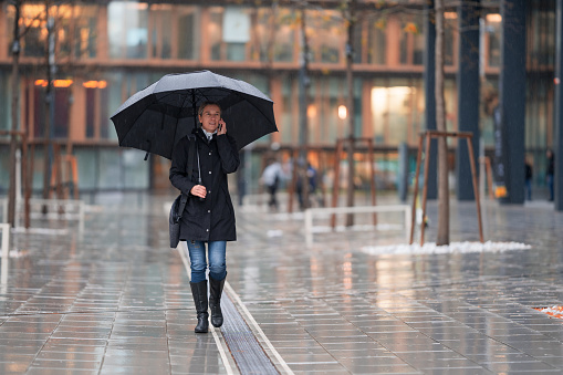 friendly 45 years old casual dressed businesswoman with huge umbrella walking through rain in urban business office district in european city on cold, windy rainy autumn day, talking on mobile phone, shallow focus background blurred