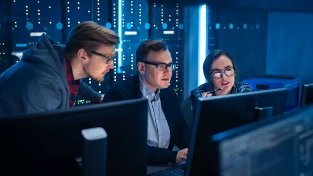 Photo of Three IT Engineers / Programmers Talking about Work, Using Computers. Technical Department with Data Server Racks. Software Development / Code Writing / Website Design / Database Architecture