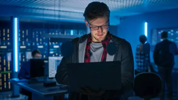 Portrait of a Smart Focused Young Man Wearing Glasses Holds Laptop. In the Background Technical Department Office with Specialists Working and Functional Data Server Racks