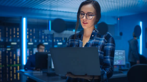 ritratto di una giovane donna intelligente che indossa occhiali tiene il laptop. nell'ufficio del reparto tecnico di base con specialisti che lavorano e rack di server dati funzionali - data center network server computer network foto e immagini stock