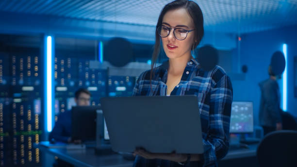 Portrait of a Smart Young Woman Wearing Glasses Holds Laptop. In the Background Technical Department Office with Specialists Working and Functional Data Server Racks Portrait of a Smart Young Woman Wearing Glasses Holds Laptop. In the Background Technical Department Office with Specialists Working and Functional Data Server Racks blockchain technology stock pictures, royalty-free photos & images