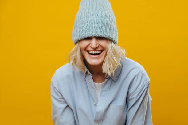 Funny blonde woman with a blue watch cap over her eyes over yellow background. Funny laughing blonde woman in a jeans shirt over yellow background. She has a blue knitted watch cap almost covering her eyes. Half length. hair covering eyes stock pictures, royalty-free photos & images