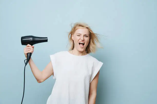 Photo of Playful blonde woman blowing herself in a face with a hairdryer over blue