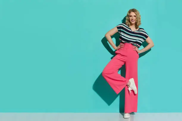 Stylish young woman in pink wide legs trousers, sneakers and striped blouse is posing on one leg. Full length studio shot on turquoise background.