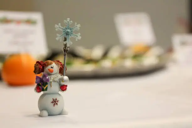 Small figure of smiling snowwoman or snowgirl holding a snowflake with copyspace. Snowman shape picture, photo or place card holder . Free space for text. Tangerine and food dish blurred on background