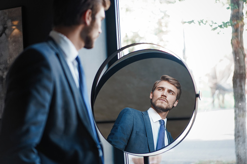 Perfect look. Reflection of handsome young man in full suit standing near the window in front of the mirror indoors