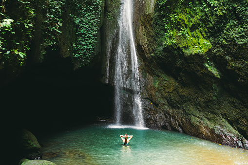 Young woman with long hair resting after hot day on Bali Island at the beautiful hidden high waterfall with green plants surroundings and turquoise lake, Indonesia