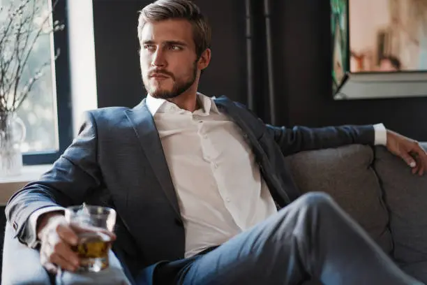 Photo of Handsome and successful businessman in stylish suit holding glass whiskey while sitting at office.