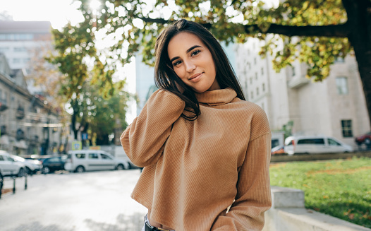 Outdoor image of beautiful young brunette woman wearing beige sweatshirt, smiling and looking directly to the camera. Pretty female student posing in the city street.